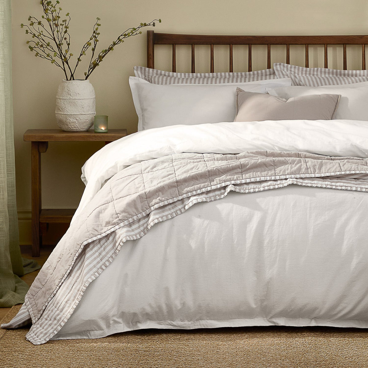Hebden Cotton Plain Duvet Cover Set in chalk white on a bed with striped pillowcases and a quilted throw, wooden headboard and nightstand in a bedroom setting.