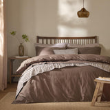 Hebden Cotton Plain Duvet Cover Set in cocoa brown on a bed with striped trim, styled in a bedroom with a dark wooden slatted headboard and amber pendant light.
