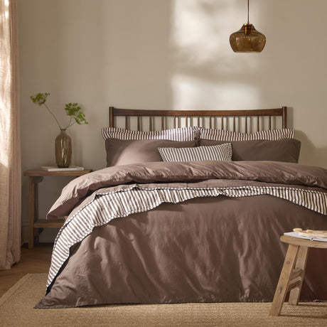 Hebden Cotton Plain Duvet Cover Set in cocoa brown on a bed with striped trim, styled in a bedroom with a dark wooden slatted headboard and amber pendant light.