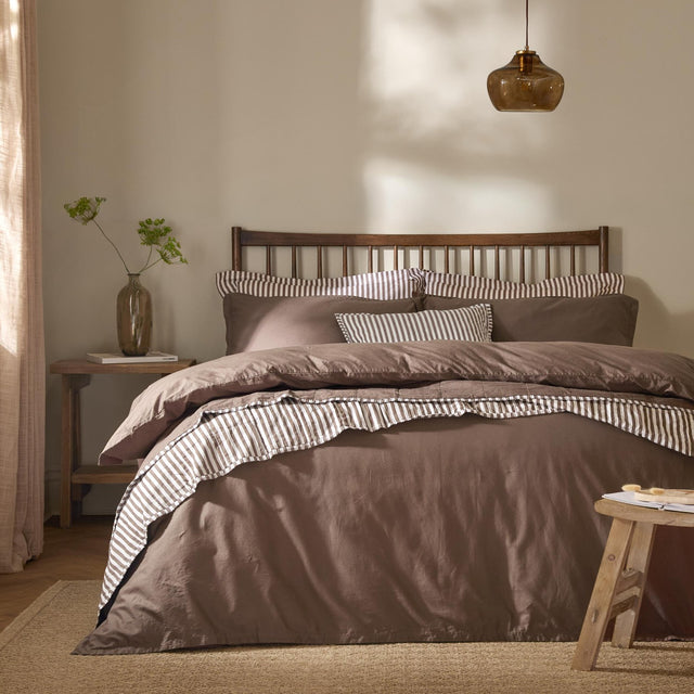 Hebden Cotton Plain Duvet Cover Set in cocoa brown on a bed with striped trim, styled in a bedroom with a dark wooden slatted headboard and amber pendant light.