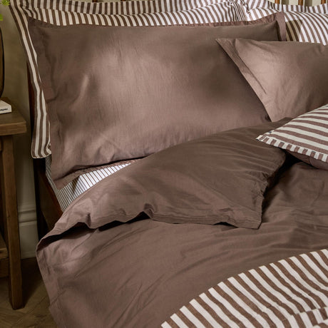 Hebden Cotton Plain Duvet Cover Set in cocoa brown on a bed, with matching pillowcases and a striped fabric border visible beneath the bedding.