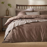 Hebden Cotton Plain Duvet Cover Set in cocoa brown on a wooden spindle bed, with striped reverse edging visible and matching pillowcases in a bedroom setting.