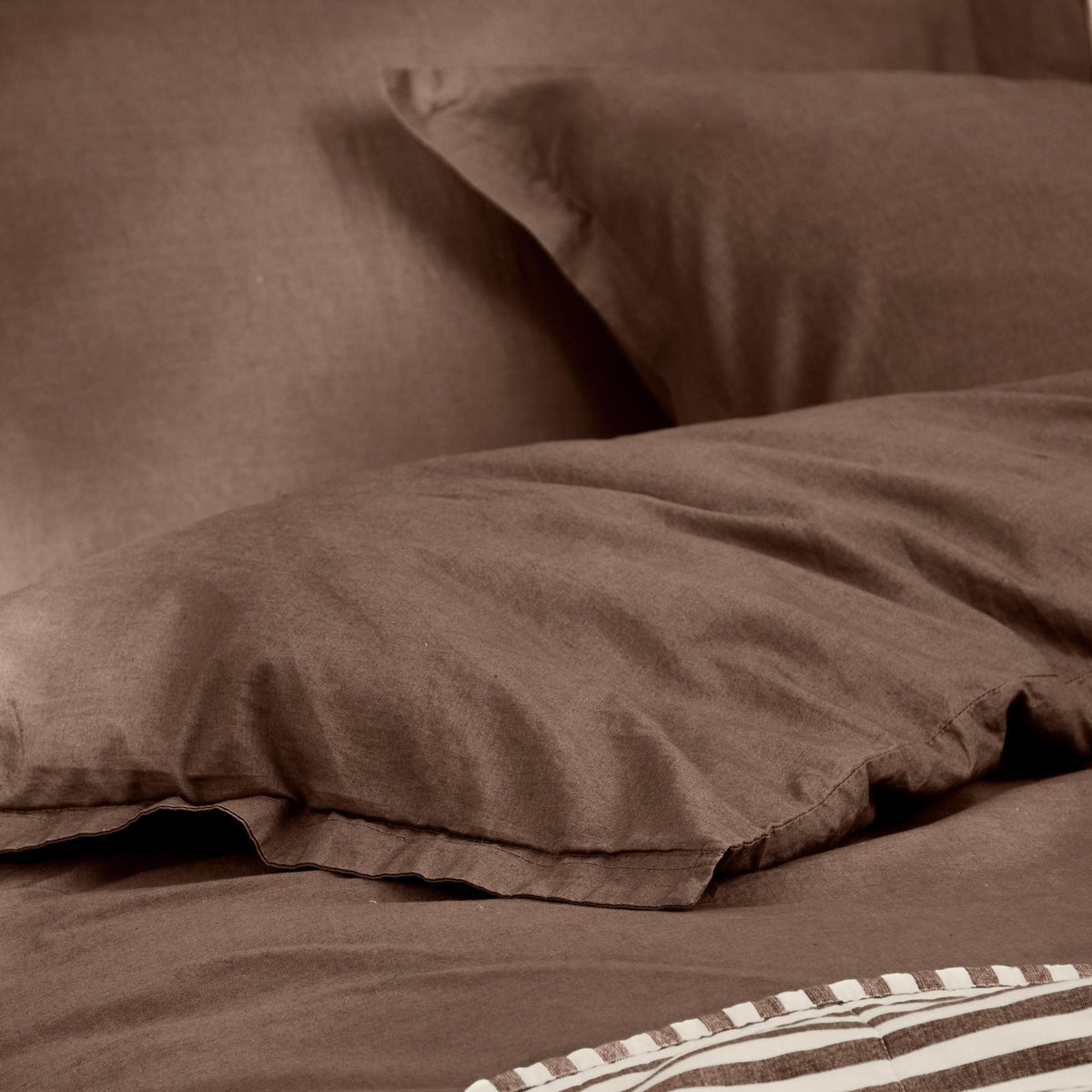 Close-up of the Hebden Cotton Plain Duvet Cover Set in cocoa brown, showing plain 100% cotton percale fabric with softly creased pillowcases and duvet cover, striped bedding visible at the edge.