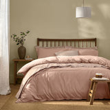 Hebden Cotton Plain Duvet Cover Set in Cotta dusky pink on a bed with striped reverse and pillowcases, wooden headboard, striped pendant shade, and bedside stool in a neutral bedroom.