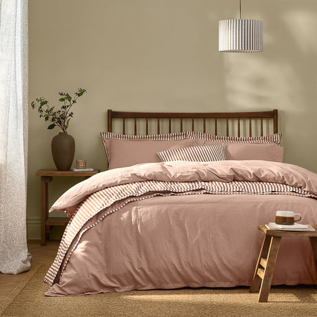 Hebden Cotton Plain Duvet Cover Set in Cotta dusky pink on a bed with striped reverse and pillowcases, wooden headboard, striped pendant shade, and bedside stool in a neutral bedroom.