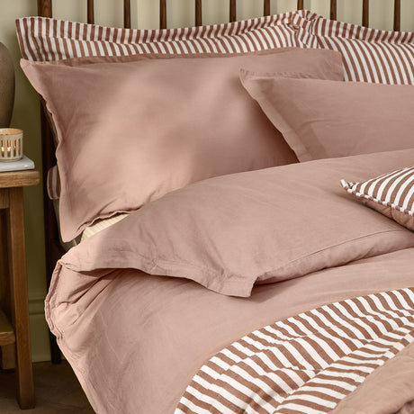 Hebden Cotton Plain Duvet Cover Set in dusky pink Cotta, with matching pillowcases and reverse tan-and-cream stripe detail, shown on a bed in a bedroom setting.