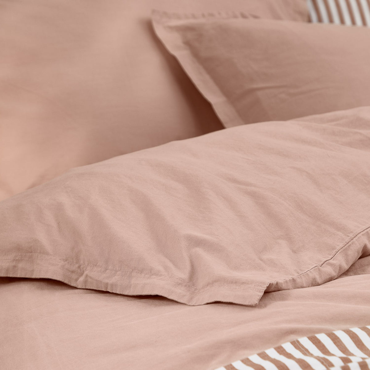 Close-up of the Hebden Cotton Plain Duvet Cover Set in Cotta, showing dusky pink washed cotton with a tan and cream striped reverse peeking through the folds.