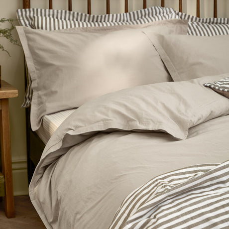 Hebden Cotton Plain Duvet Cover Set in Stone, a grey-beige cotton duvet cover with matching pillowcases, shown on a bed with striped accent bedding in a bedroom setting.