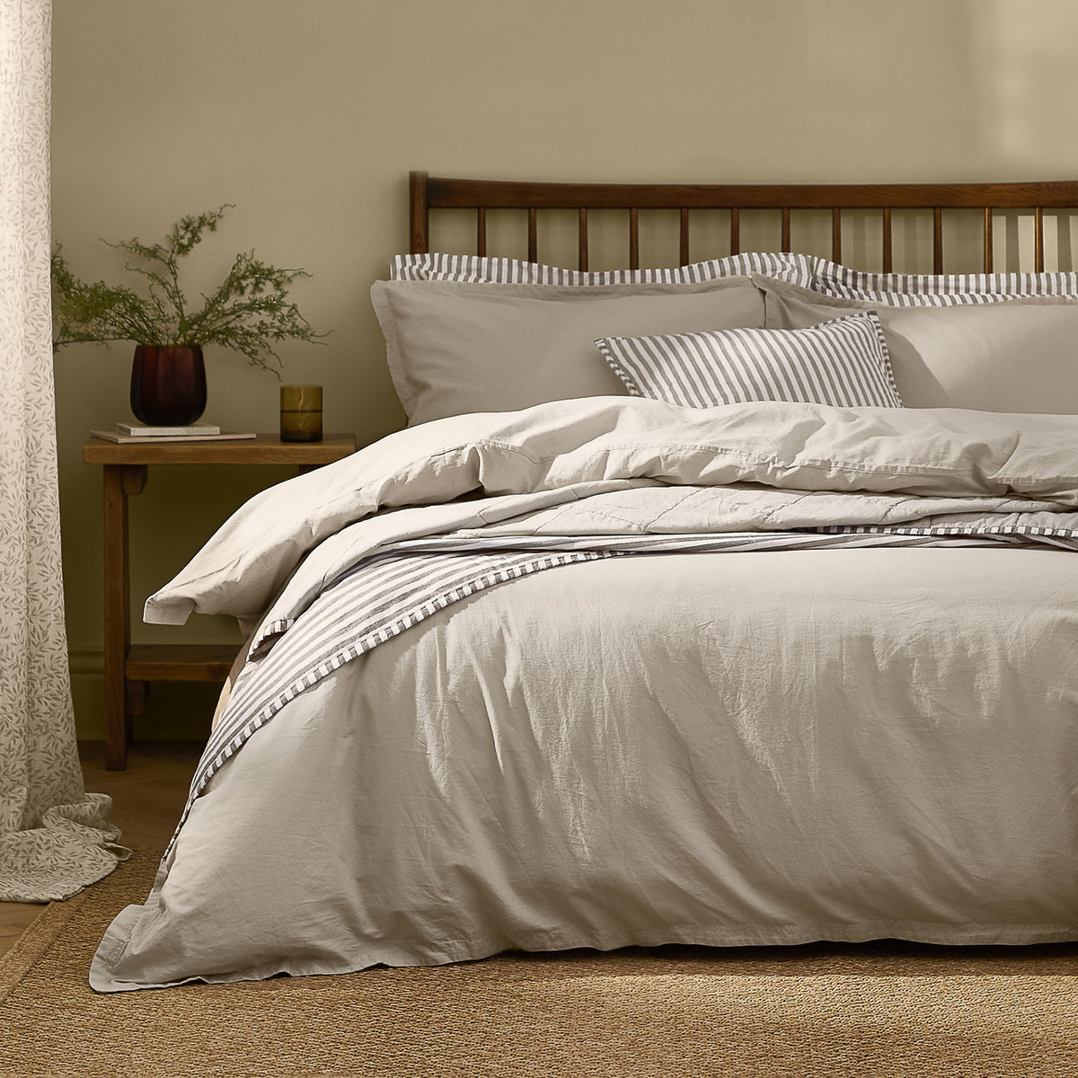 Hebden Cotton Plain Duvet Cover Set in Stone on a wooden spindle bed, styled with striped cushions in a bedroom setting.