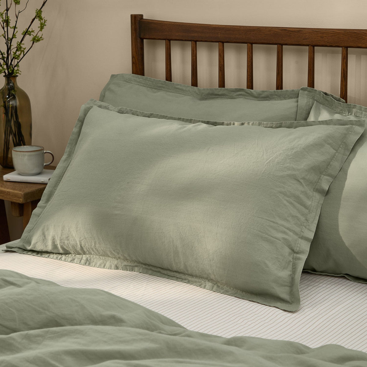 Hebden Percale Cotton Pillowcase Pair in Bay Green, shown with flanged edges on a bedroom bed against a dark wood headboard.