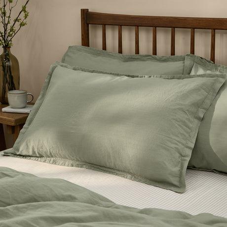 Hebden Percale Cotton Pillowcase Pair in Bay Green, shown with flanged edges on a bedroom bed against a dark wood headboard.