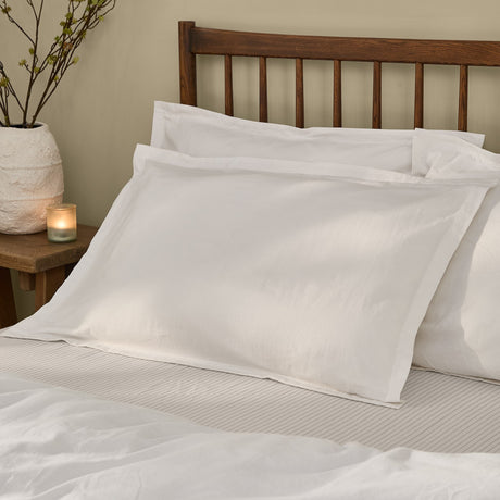 Hebden Percale Cotton Pillowcase Pair in Chalk, styled on a bed with a wooden headboard, showing the plain-weave white cotton finish and folded flap detail.