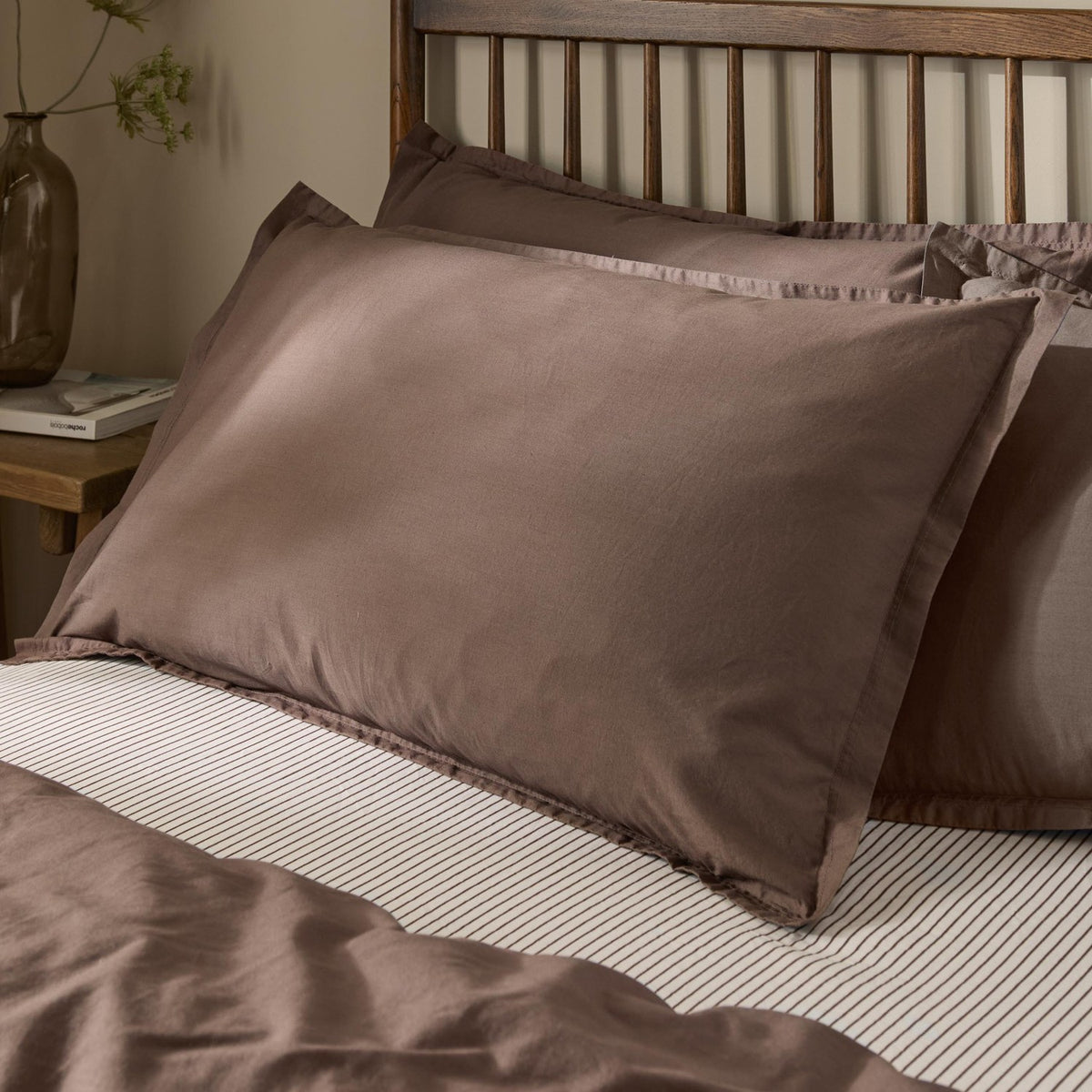 Hebden Percale Cotton Pillowcase Pair in Cocoa Brown, styled on a bed with striped linen against a wooden slatted headboard.