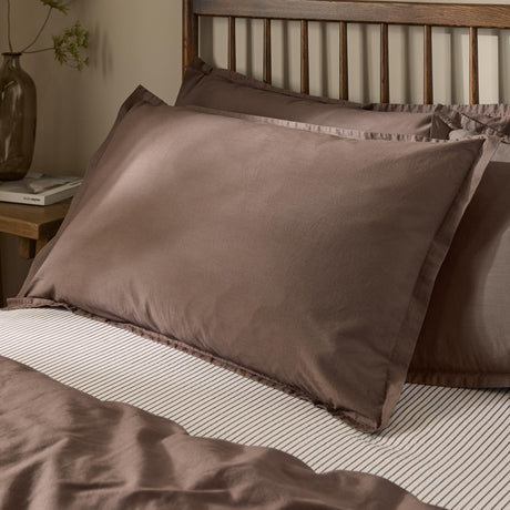 Hebden Percale Cotton Pillowcase Pair in Cocoa Brown, styled on a bed with striped linen against a wooden slatted headboard.