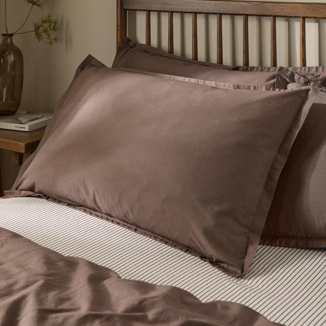 Hebden Percale Cotton Pillowcase Pair in Cocoa Brown, styled on a bed with striped linen against a wooden slatted headboard.