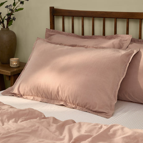 Hebden Percale Cotton Pillowcase Pair in Cotta pink, styled on a bed with a dark wood headboard, showing the cotton percale finish and stitched edge detail.