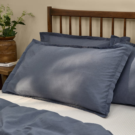 Hebden Percale Cotton Pillowcase Pair in Denim Blue, shown styled on a bed with matching duvet cover against a wooden headboard in a bedroom setting.