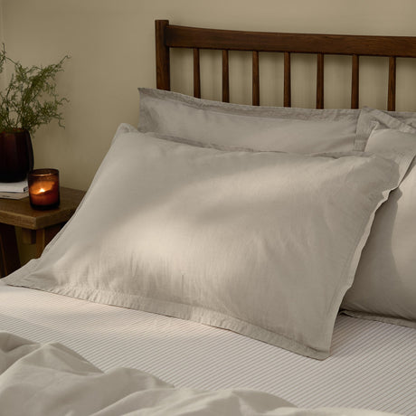 Hebden Percale Cotton Pillowcase Pair in stone, styled on a bed against a dark wooden headboard with a bedside candle and plant in a softly lit bedroom.