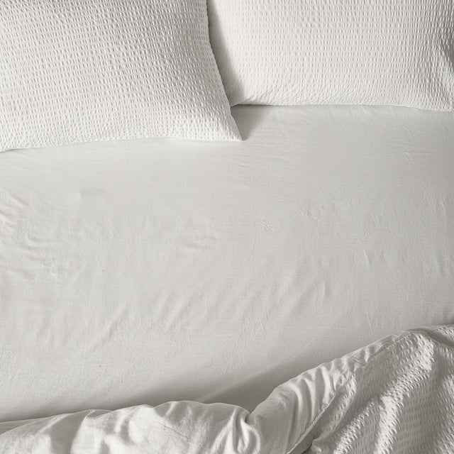 Hebden Percale Cotton Fitted Sheet in Chalk, a soft off-white 100% cotton bedsheet with a crinkled matt texture, shown fitted on a bed with matching waffle-textured pillowcases.