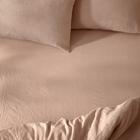 Hebden Percale Cotton Fitted Sheet in Cotta pink, shown on a bed with waffle-textured pillows and a matching quilted cover in soft natural light.