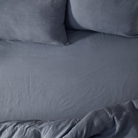 Hebden Percale Cotton Fitted Sheet in denim blue, shown fitted on a bed with matching waffle-textured pillows and a folded corner of throw fabric.