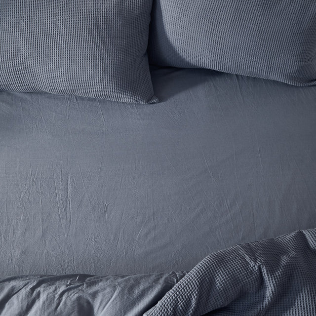 Hebden Percale Cotton Fitted Sheet in denim blue, shown fitted on a bed with matching waffle-textured pillows and a folded corner of throw fabric.
