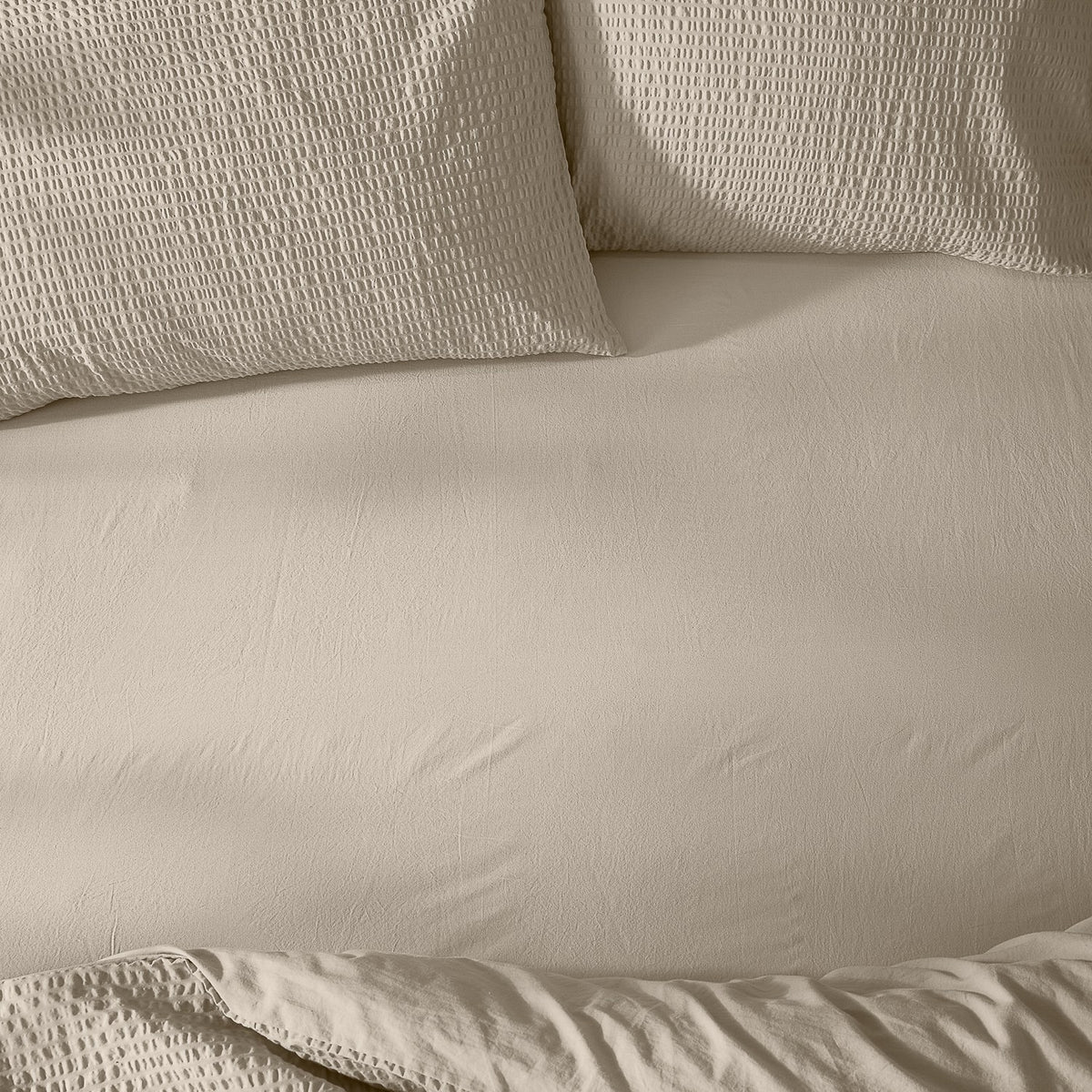 Hebden Percale Cotton Fitted Sheet in stone, shown smoothly fitted on a bed with matching waffle-textured pillowcases in the same warm beige tone.