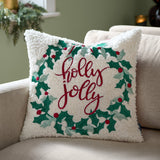 Square 45x45cm bouclé cushion with embroidered holly wreath and bow in green and red, "holly jolly" lettering, shown on a beige sofa.
