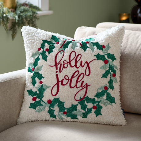 Square 45x45cm bouclé cushion with embroidered holly wreath and bow in green and red, "holly jolly" lettering, shown on a beige sofa.