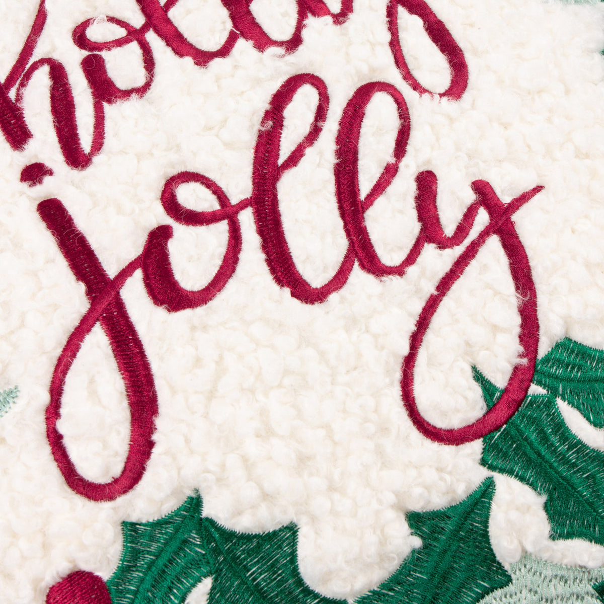 Close-up of the Holly Wreath Bow Velvet Cushion showing "Holly Jolly" embroidered in red script on textured white bouclé, framed by green holly leaves.
