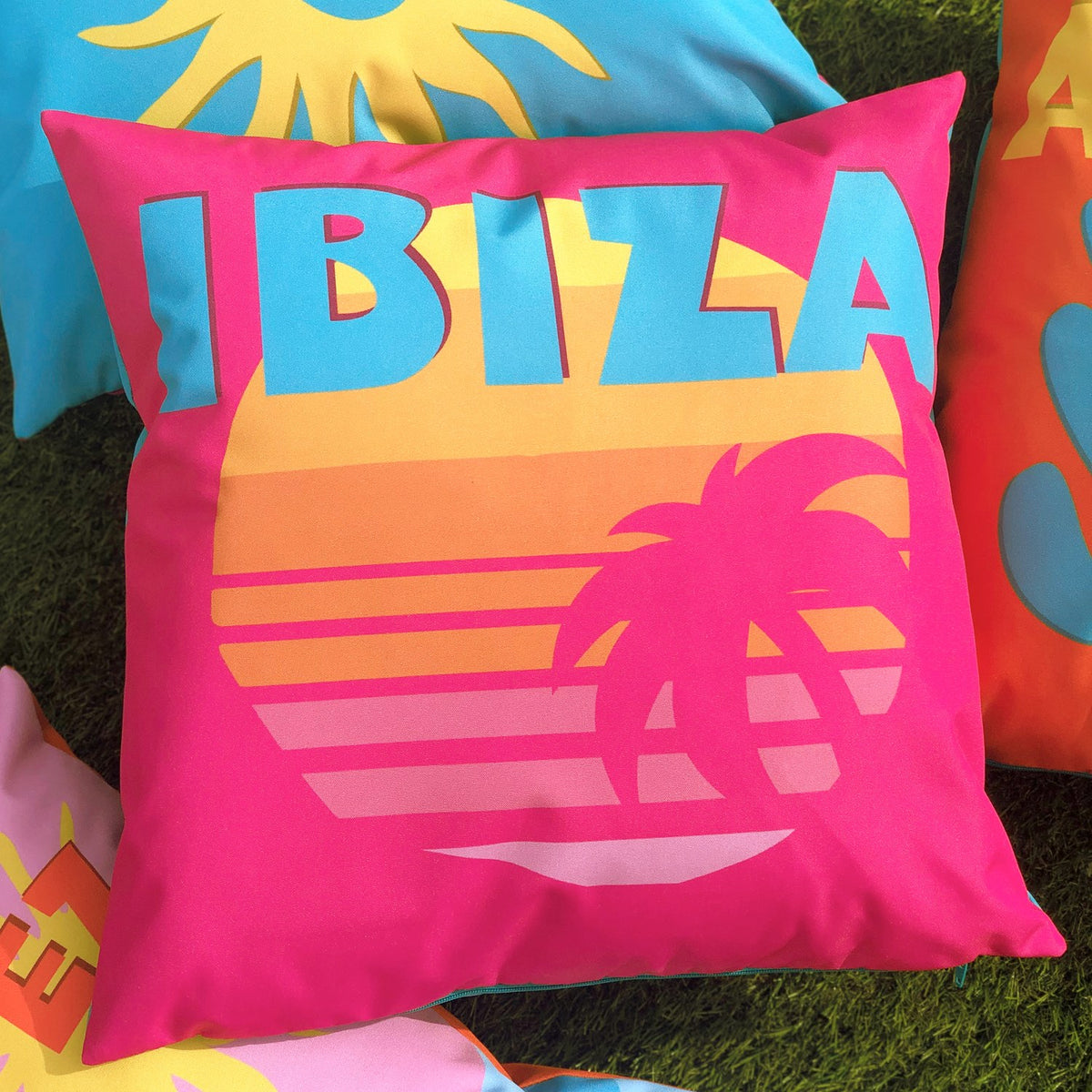 Square outdoor cushion in hot pink polyester with "IBIZA" in blue lettering above a striped sunset and palm tree graphic, shown among other patterned cushions on grass.