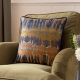 Inca Jacquard Velvet Cushion in royal blue, gold and orange abstract woven pattern with navy velvet piping, shown on an olive green armchair.