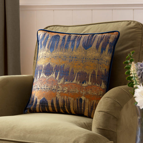 Inca Jacquard Velvet Cushion in royal blue, gold and orange abstract woven pattern with navy velvet piping, shown on an olive green armchair.