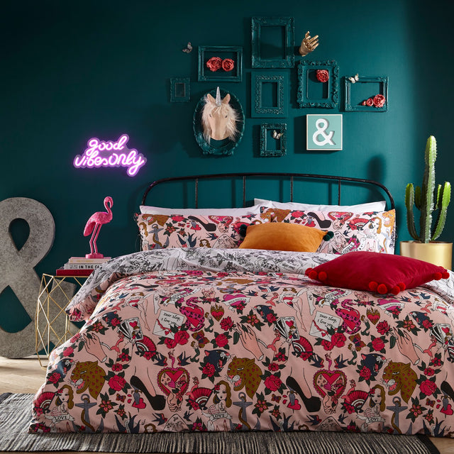 Blush pink Inked Reversible Duvet Cover Set with tattoo-style roses, hearts, pin-up figures and love-letter motifs, styled on a bed in a teal bedroom with orange and red cushions.