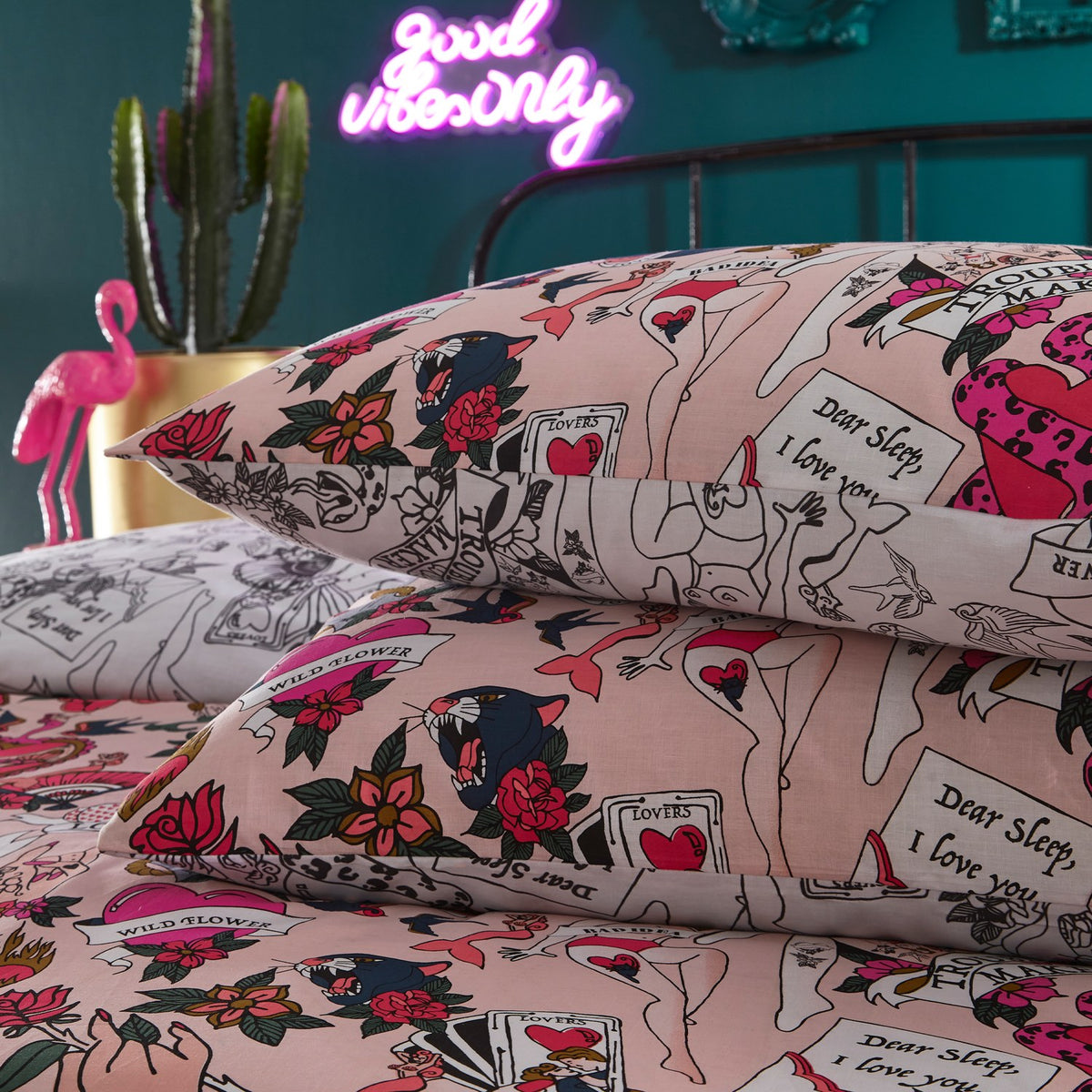 Reversible Blush Abstract Duvet Cover Set on a bed with tattoo-style prints of hearts, roses, pin-up figures and a Trouble Maker banner, in a bedroom with neon sign backdrop.