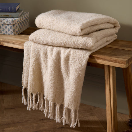 Isla Plain Polyester Throw in cream, folded with a textured woven surface and twisted tassel fringe, draped over a wooden bench beside stacked books.