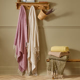 Isla Plain Polyester Throw in cream, woven with fringed tassel ends, hanging on a wooden wall-mounted hook alongside a pink throw and folded stacked throws on a bench.