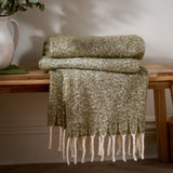 Isla Plain Polyester Throw in green folded on a wooden bench, showing its woven two-tone texture and cream tasselled fringe draping down.