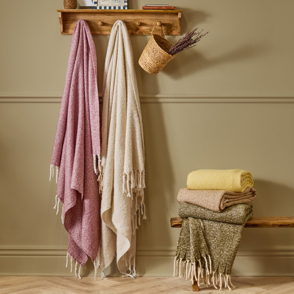 Isla Plain Polyester Throw in green folded with cream and yellow throws, tasselled fringing visible, styled on a wooden bench beside hanging pink and cream throws.