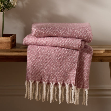 Isla Plain Polyester Throw in Heather, a dusky pink woven blanket with flecked texture and cream tasselled fringe, folded on a wooden bench.