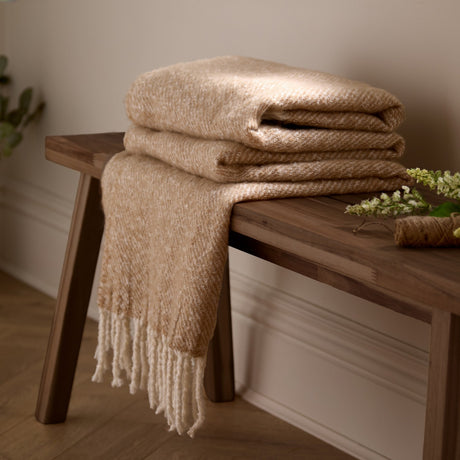 Isla Plain Polyester Throw in natural beige, folded on a wooden bench with tasselled fringe draped down, showing a fine diagonal woven texture.