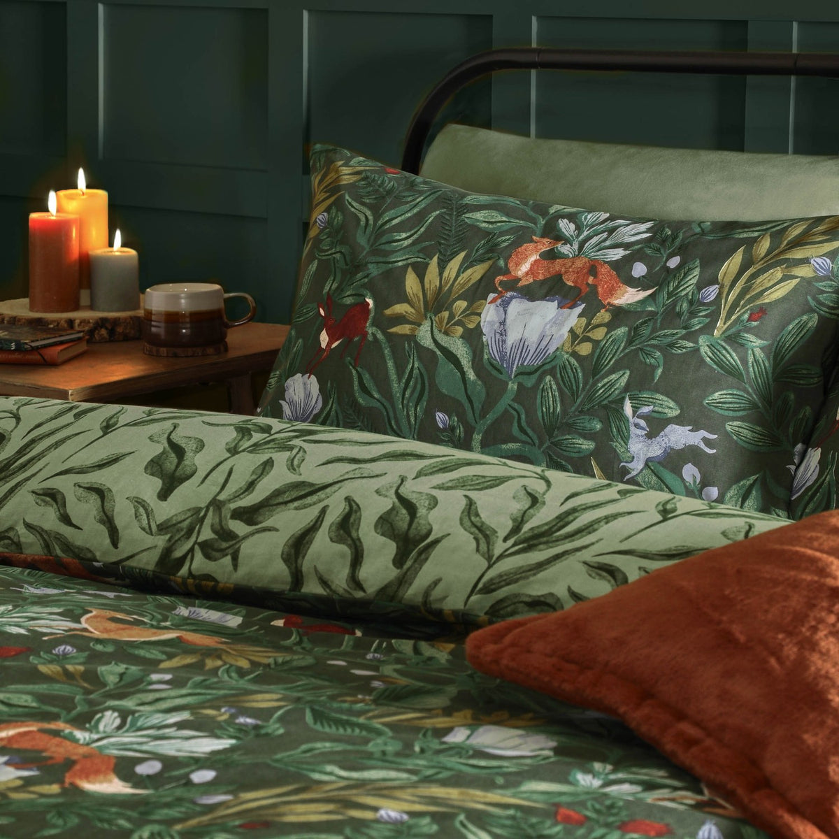 Forest green Into The Woods reversible duvet set with woodland-print pillowcase featuring foxes, hares and foliage, styled on a bed with rust cushions in a dark green bedroom.