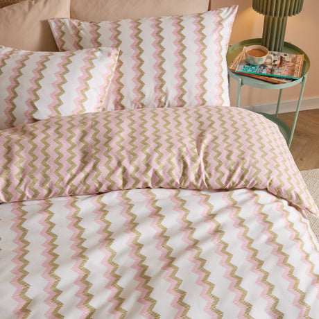 Izzie Reversible Striped Duvet Cover Set in pink and olive-green zig-zag on white, shown with matching pillowcases on a bedroom bed beside a side table.