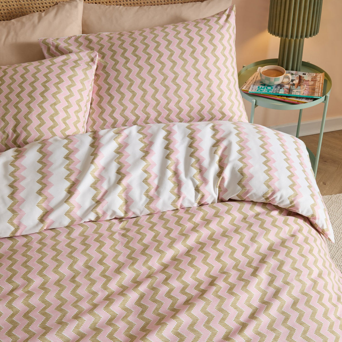 Izzie Reversible Striped Duvet Cover Set in pink and olive-green zig-zag on white, shown with matching pillowcases on a bedroom bed beside a green side table.