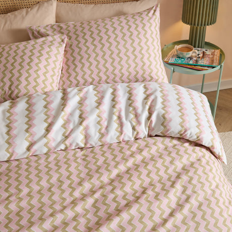 Izzie Reversible Striped Duvet Cover Set in pink and olive-green zig-zag on white, shown with matching pillowcases on a bedroom bed beside a green side table.