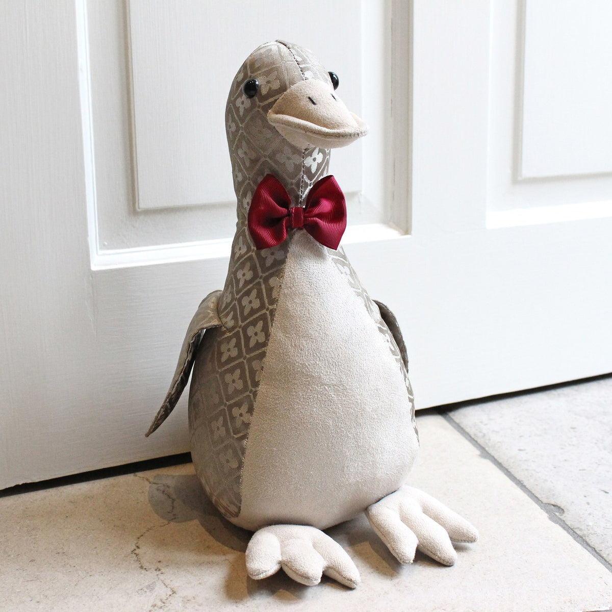 Jacquard Duck Geometric Polyester Door Stop in grey and cream, with diamond floret pattern, red bow tie and bead eyes, shown standing beside a white panelled door.