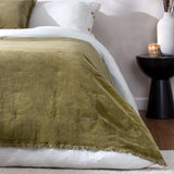 Jaye Velvet Cotton Bedspread in Moss green with fringed edge and tufted detail, layered over white bedding on a bed beside a black side table.