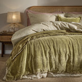 Jaye Velvet Cotton Bedspread in Moss green with fringed edge, shown layered on a bed with striped linen and cushions against a wooden cane headboard.