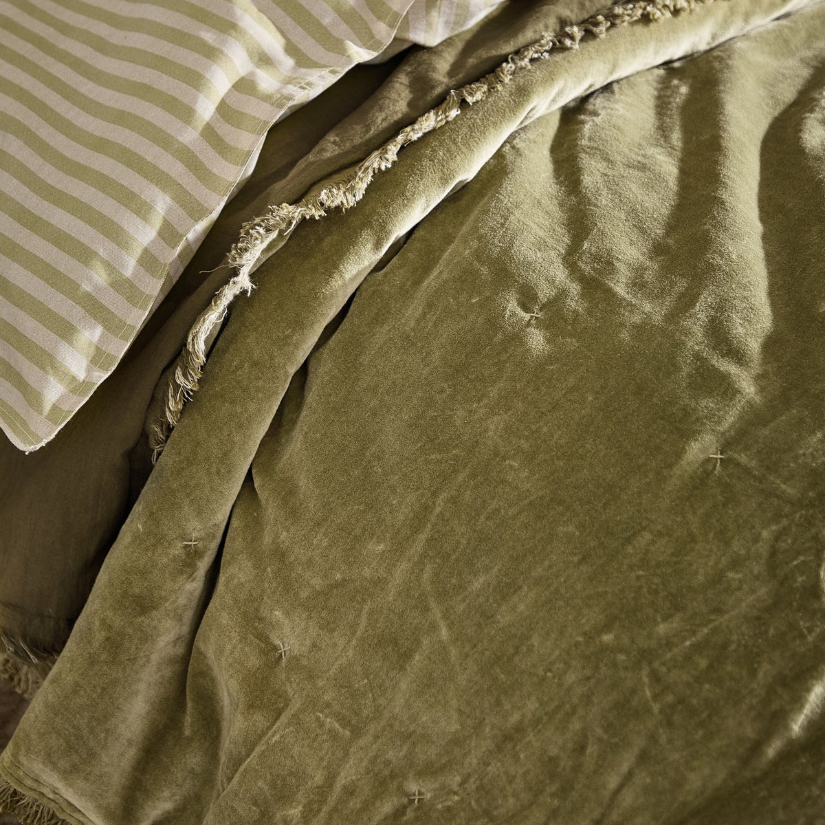 Jaye Velvet Cotton Bedspread in Moss, a soft olive-green quilted velvet throw with a frayed edge trim, shown folded on a bed beside striped pillowcases.
