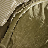 Jaye Velvet Cotton Bedspread in Moss, a soft olive-green quilted velvet throw with a frayed edge trim, shown folded on a bed beside striped pillowcases.